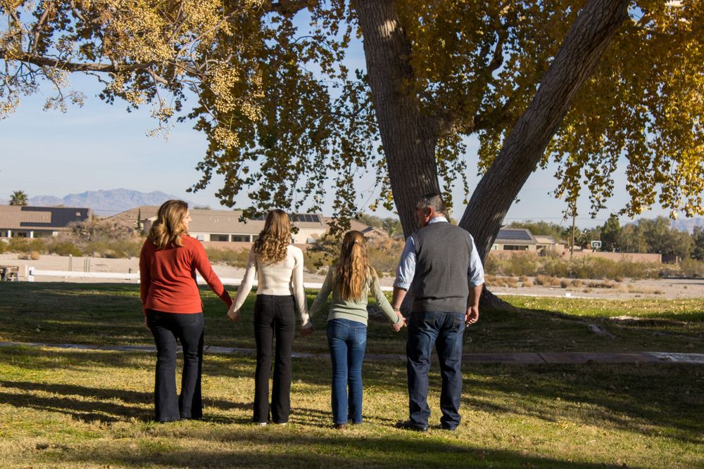 A family of four holds hands while standing under a large autumn tree in a park setting.
