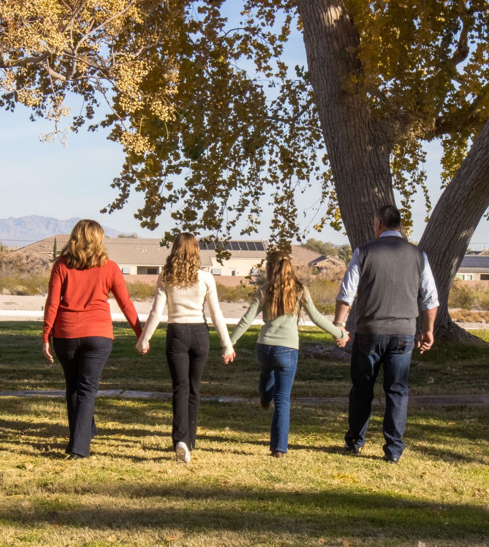 Family of four walking hand in hand across a green lawn beneath a large autumn tree.