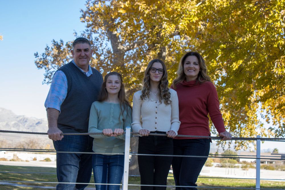Family of four smiling together at a white fence with golden autumn trees and mountains behind.