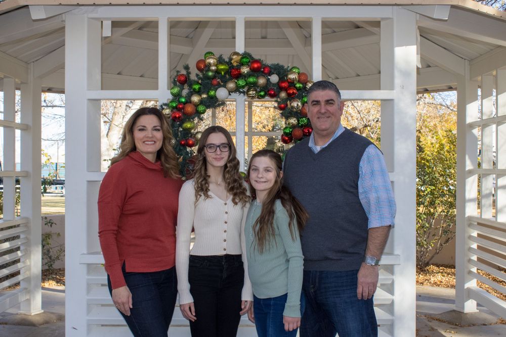 Family of four smiling together under a gazebo decorated with a colorful Christmas ornament wreath.