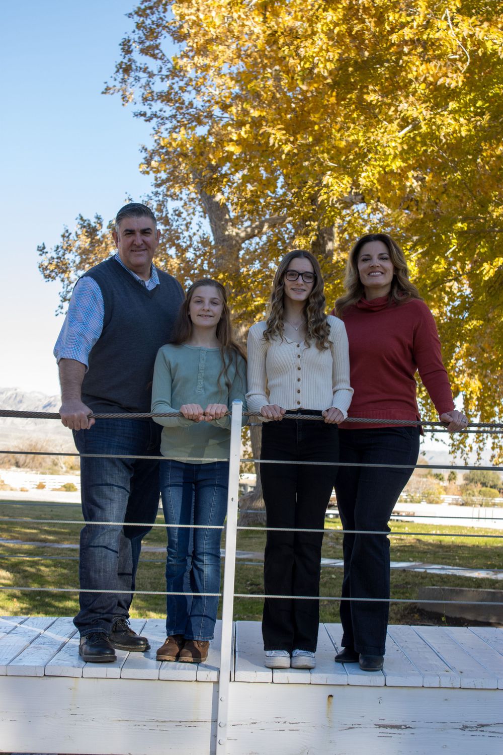 Family of four posing at a white fence with vibrant golden autumn trees in the background.
