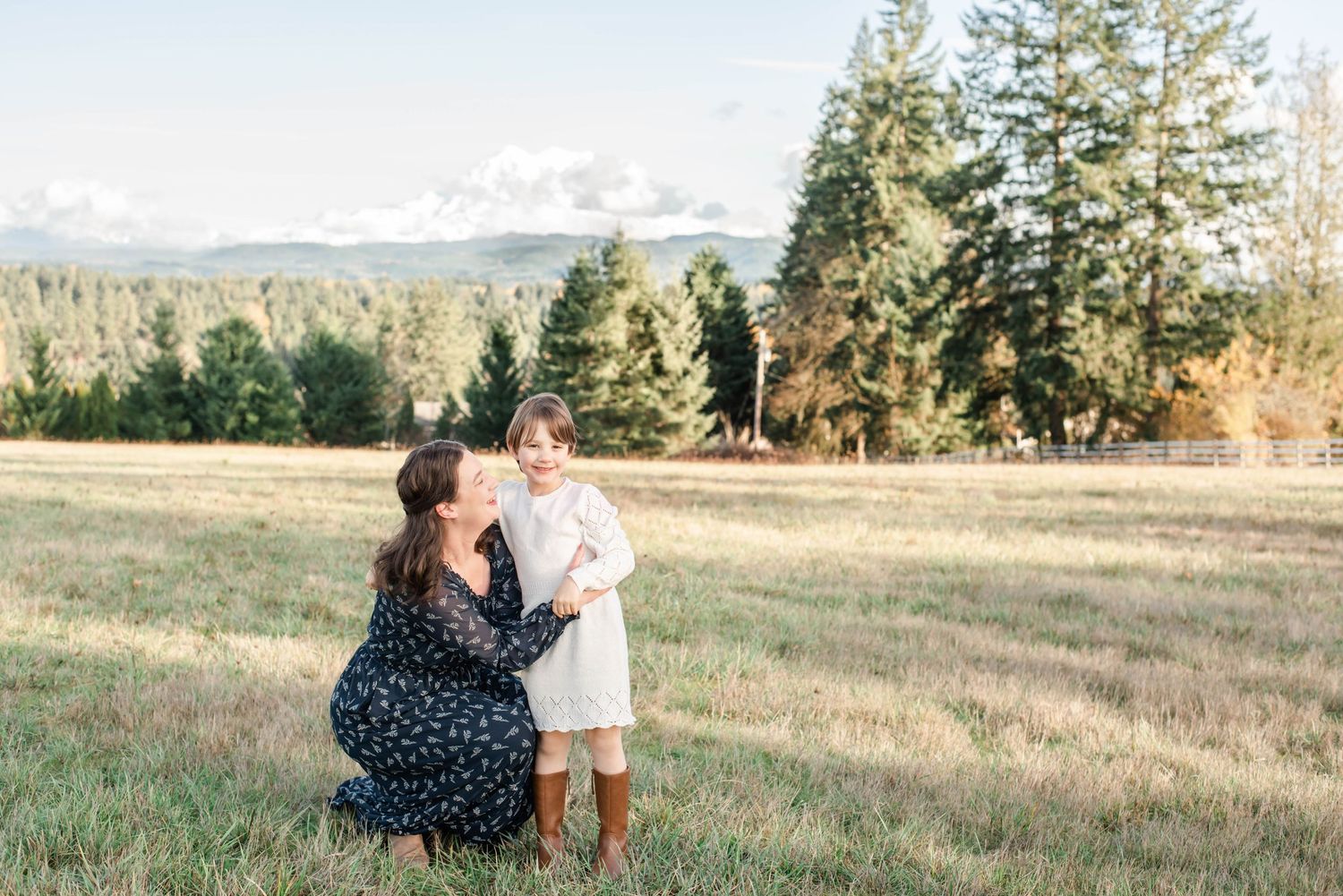 A mother and daughter share tender moments in a grassy field during golden hour with mountains in the background.