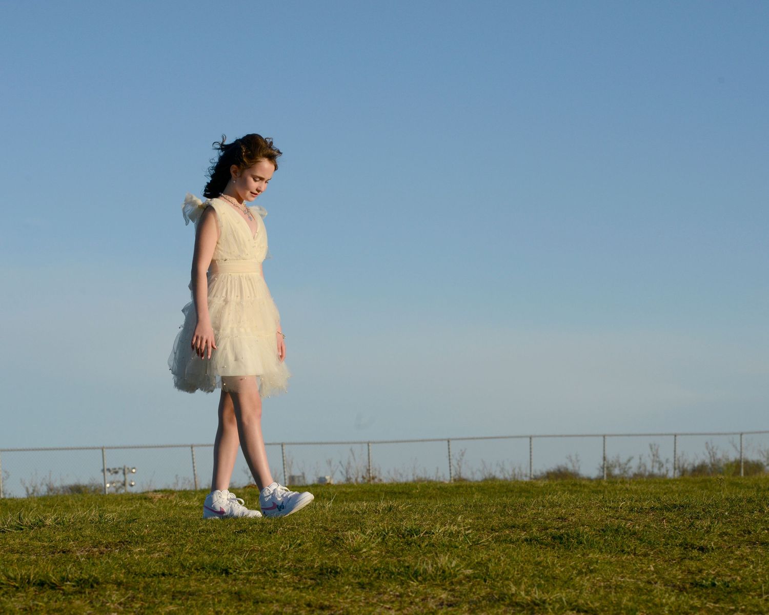 A person in a white dress and sneakers stands on a grassy hilltop against a bright blue sky.