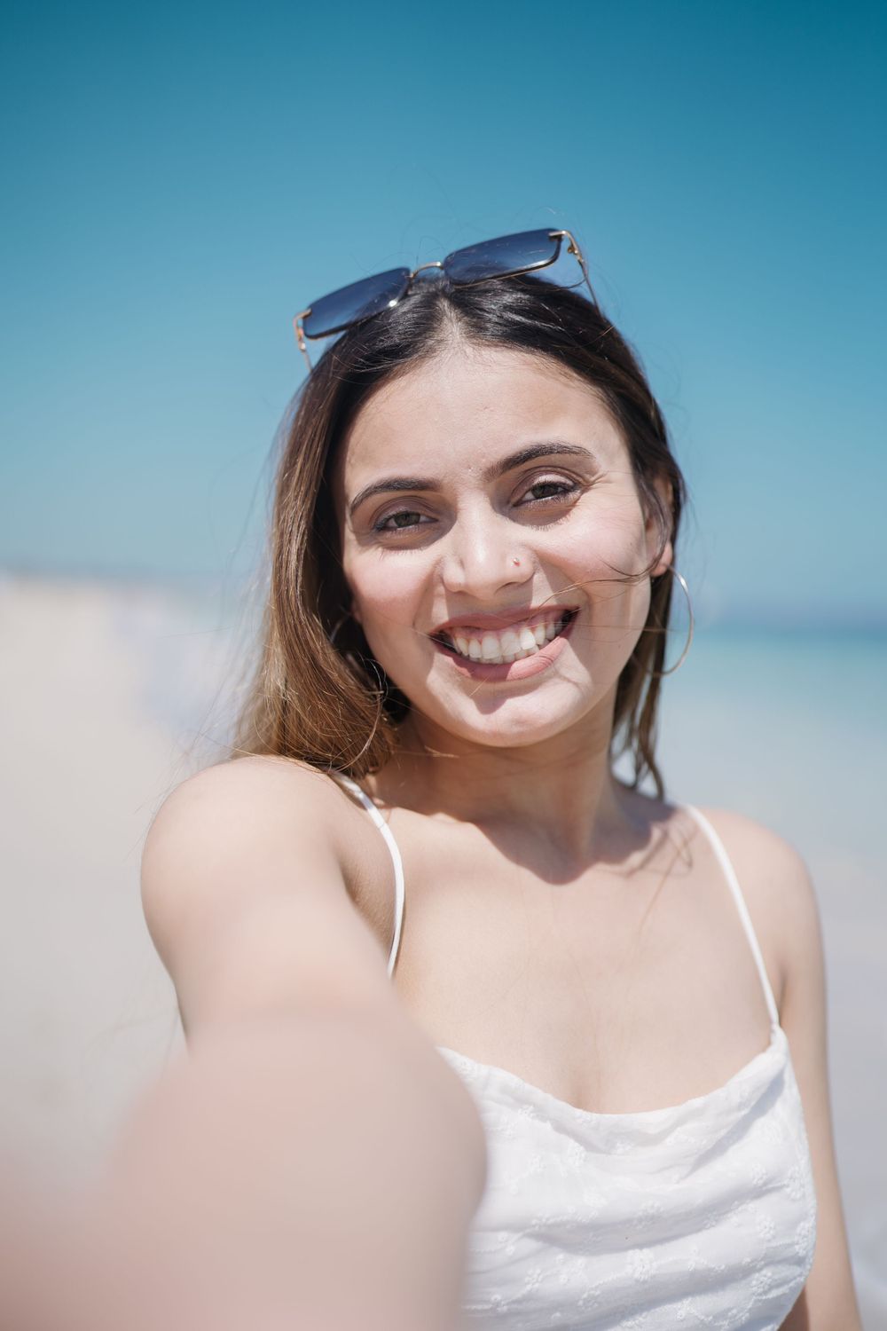 Person wearing a white tank top and sunglasses takes a selfie at the beach on a sunny day with blue skies.