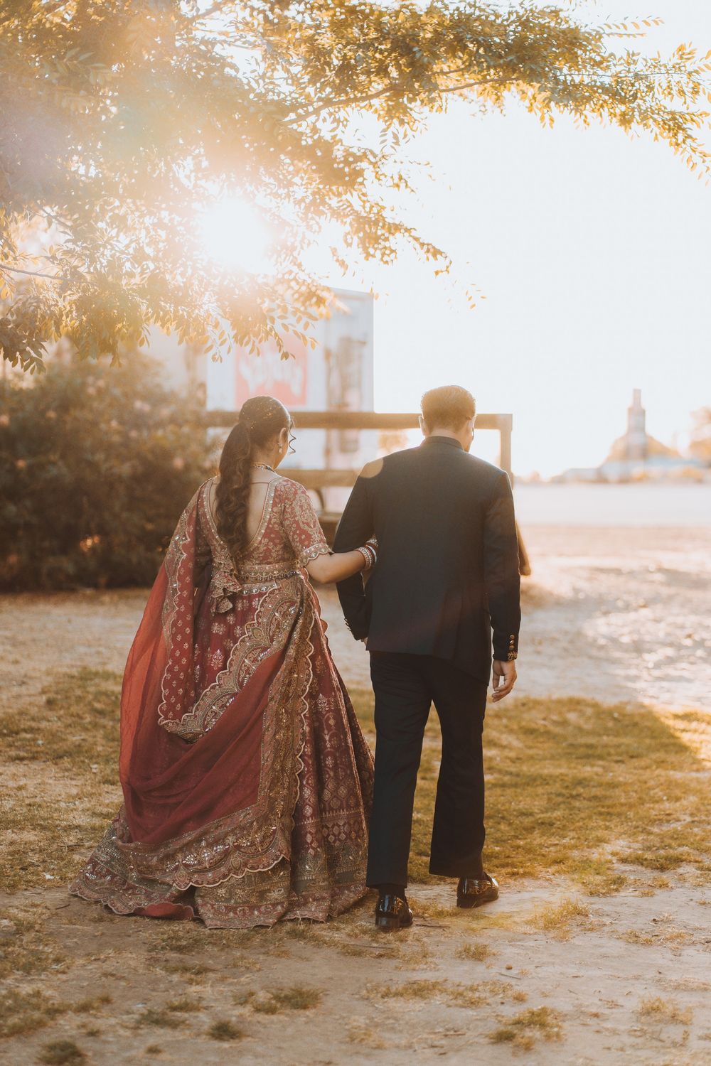 A couple in traditional South Asian wedding attire walks together during golden hour sunset in a grassy outdoor setting.