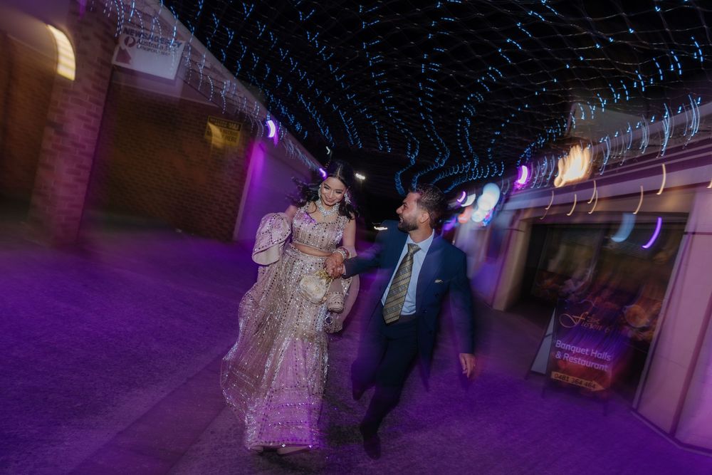 Indian bride and groom walking under glowing string lights at night, dressed in traditional wedding attire.