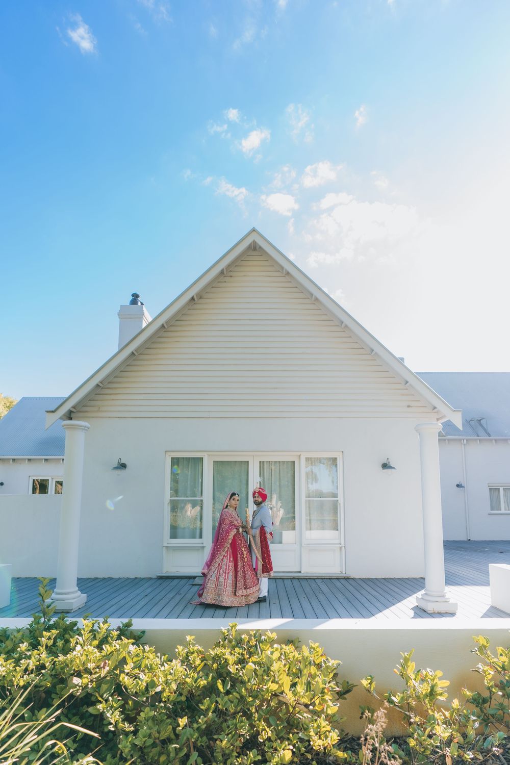 Indian bride and groom in traditional attire standing on white house porch under blue sky.