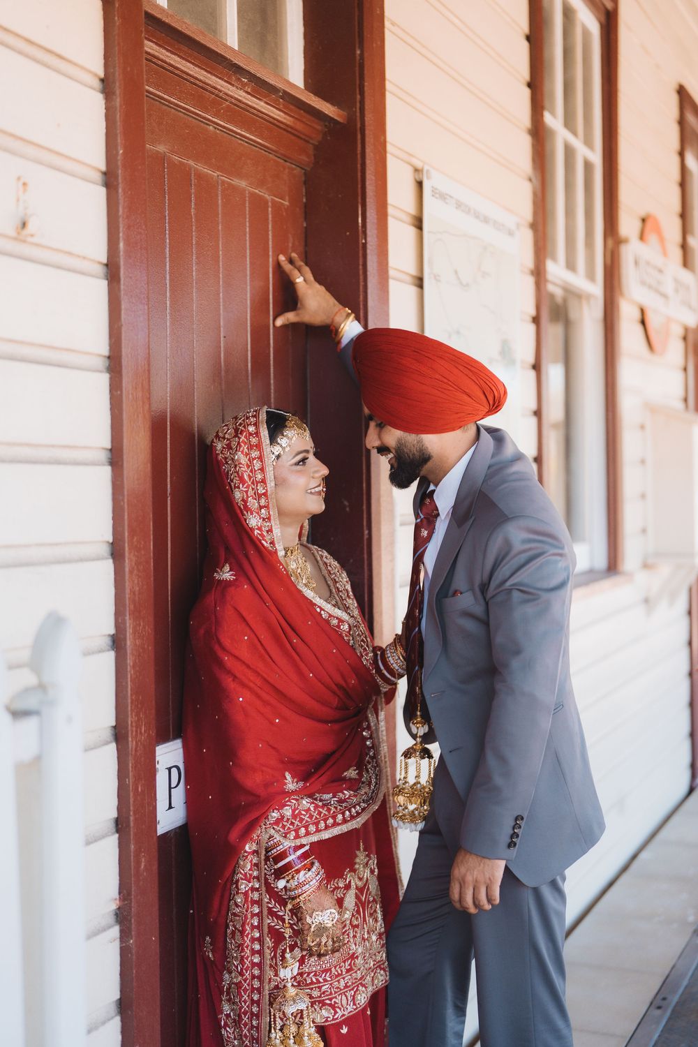 A couple in traditional wedding attire, with the bride in a red sari and groom in grey suit, pose together against a wall.
