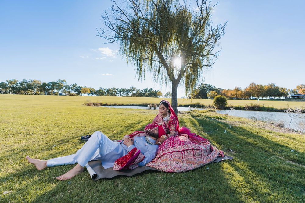 Indian couple in traditional wedding attire sitting under a weeping willow tree by a lake on a sunny day.