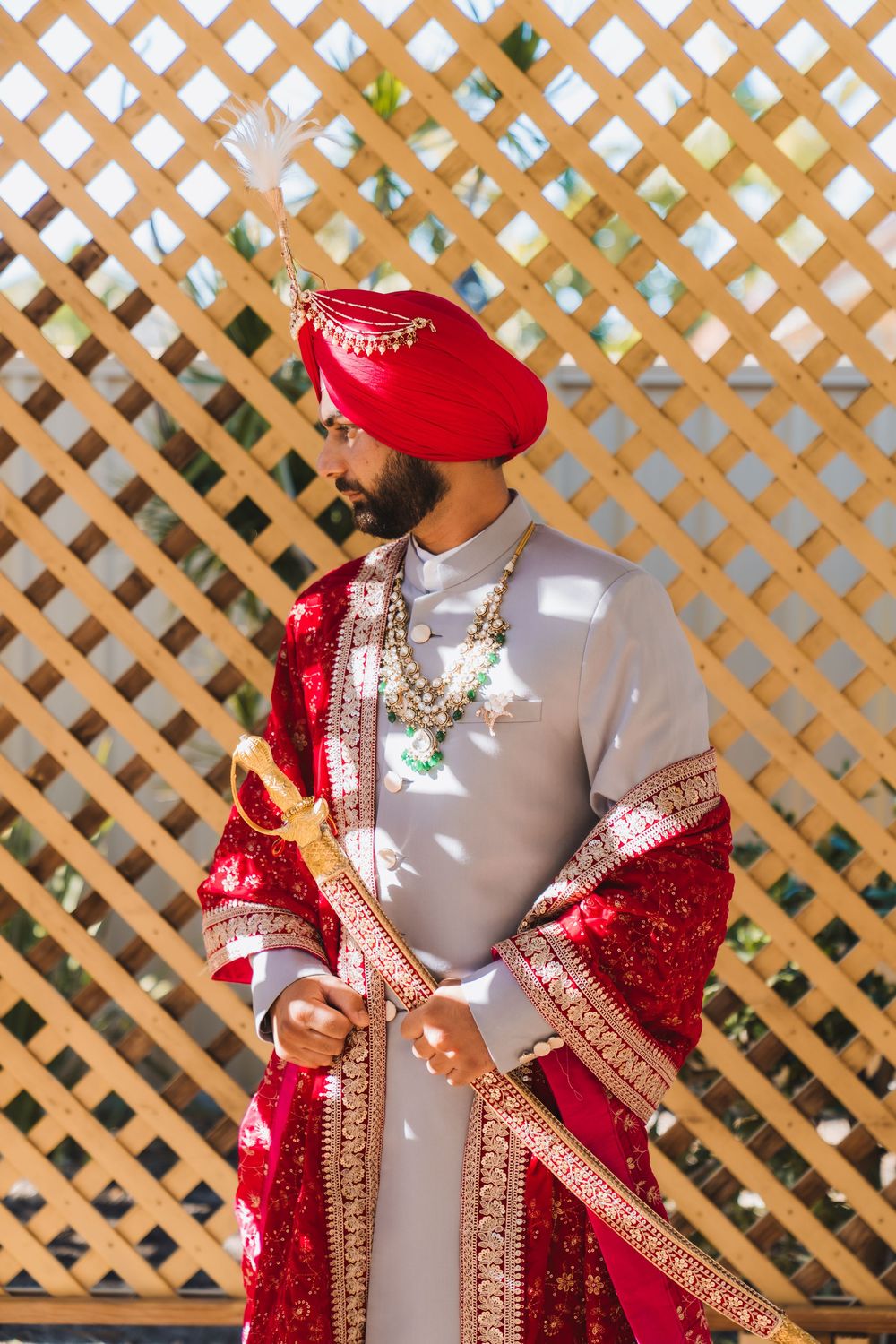 Sikh groom in traditional red sherwani, turban with kalgi, and gold jewelry poses outdoors.