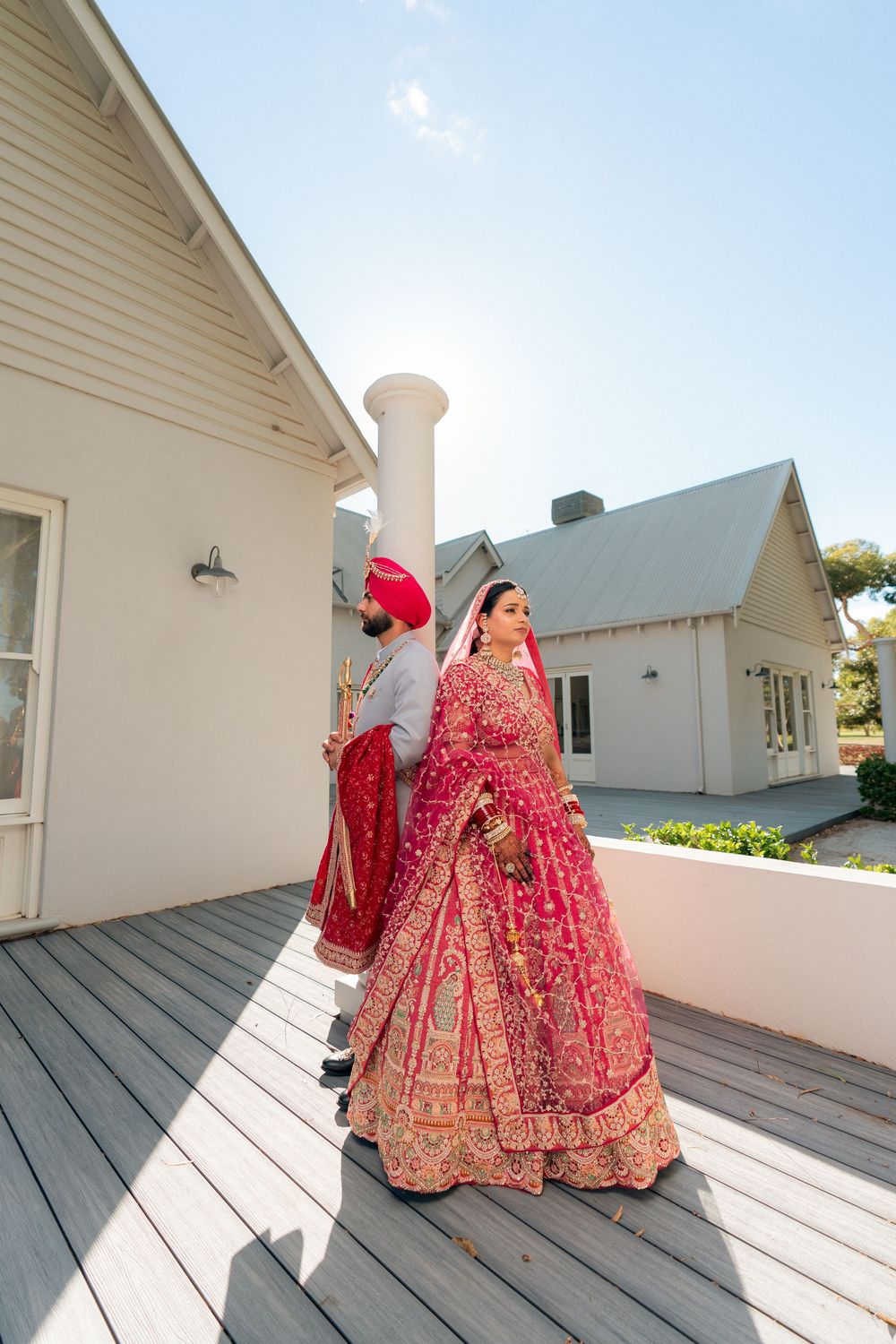 Sikh bride in red lehenga and groom in red turban walking on wooden deck outside white venue.