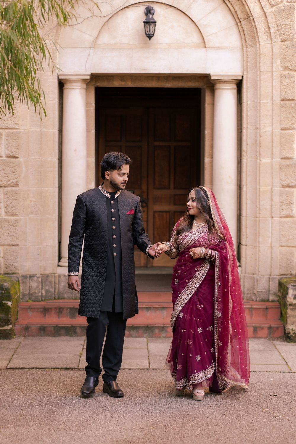 Indian couple in traditional wedding attire holding hands outside ornate building, bride in red saree, groom in black sherwani.
