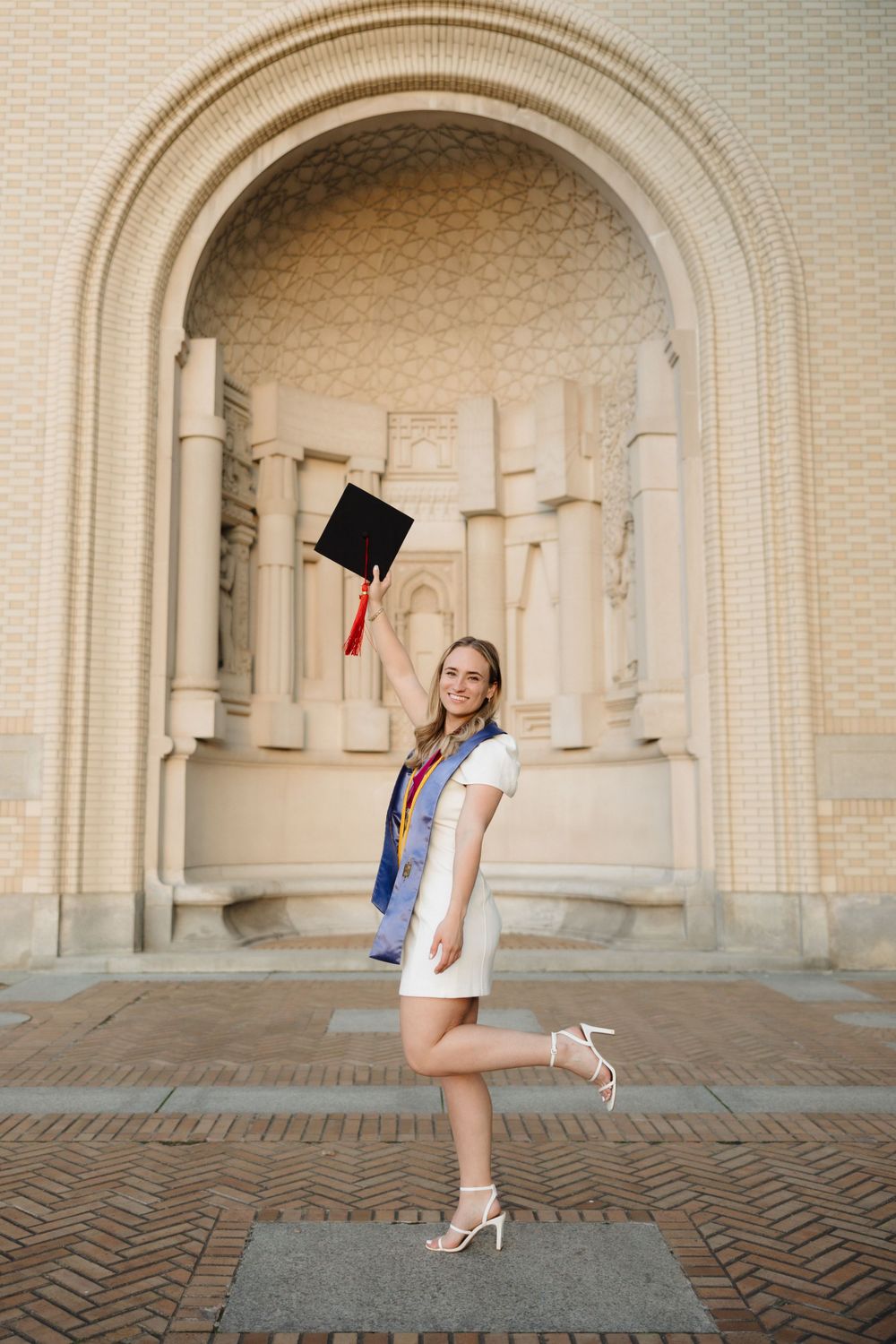 Graduate in white dress tossing cap in front of ornate archway during celebration photoshoot.