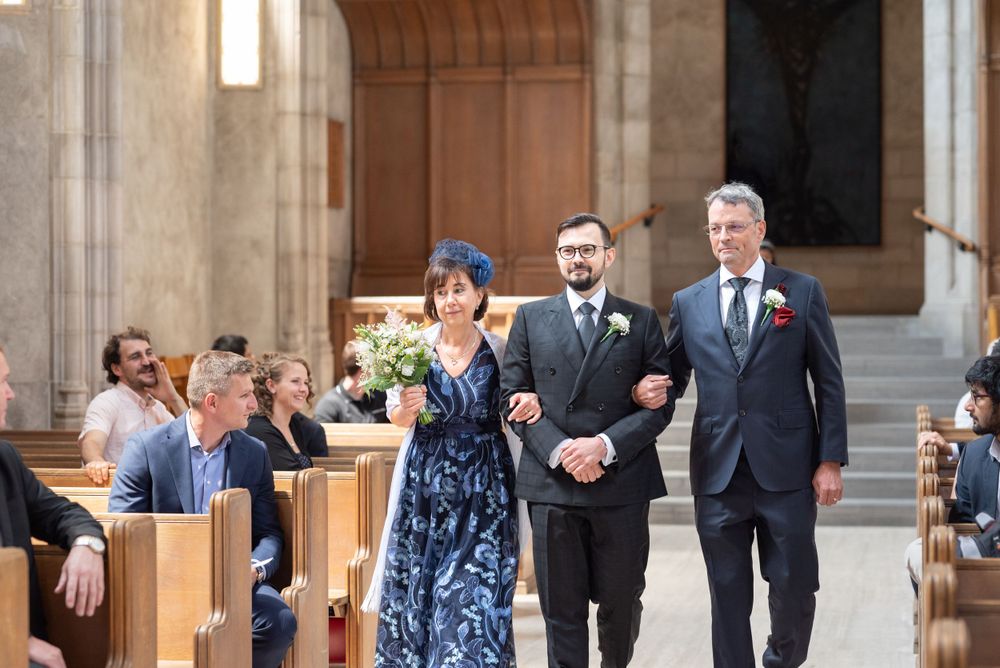 Groom walking down the aisle with his parents, as wedding party members proceed during a formal ceremony with guests seated in wooden pews.