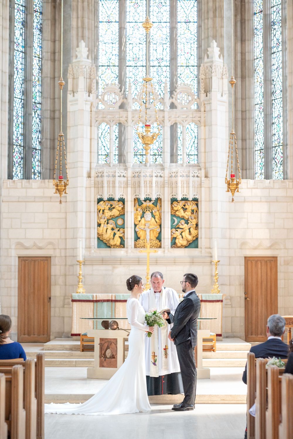 A traditional wedding ceremony at Trinity College Chapel in front of a stained glass altar with gothic architecture.