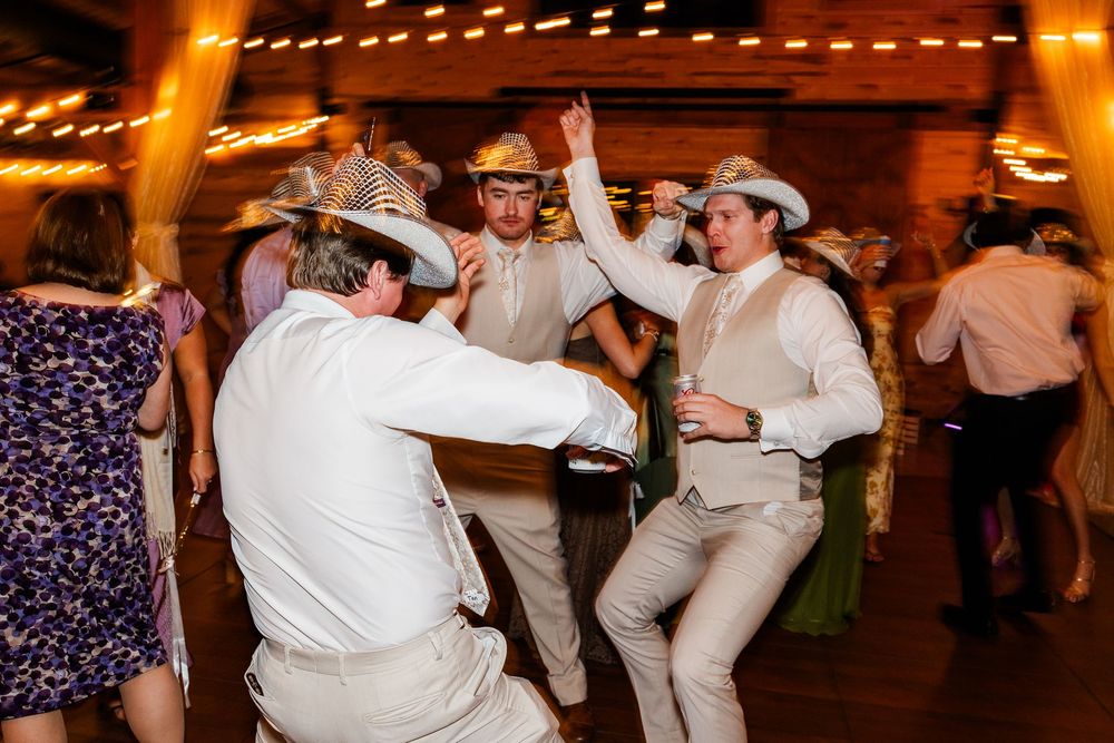 Guests in cowboy hats dancing energetically at a rustic wedding reception on a wooden dance floor.