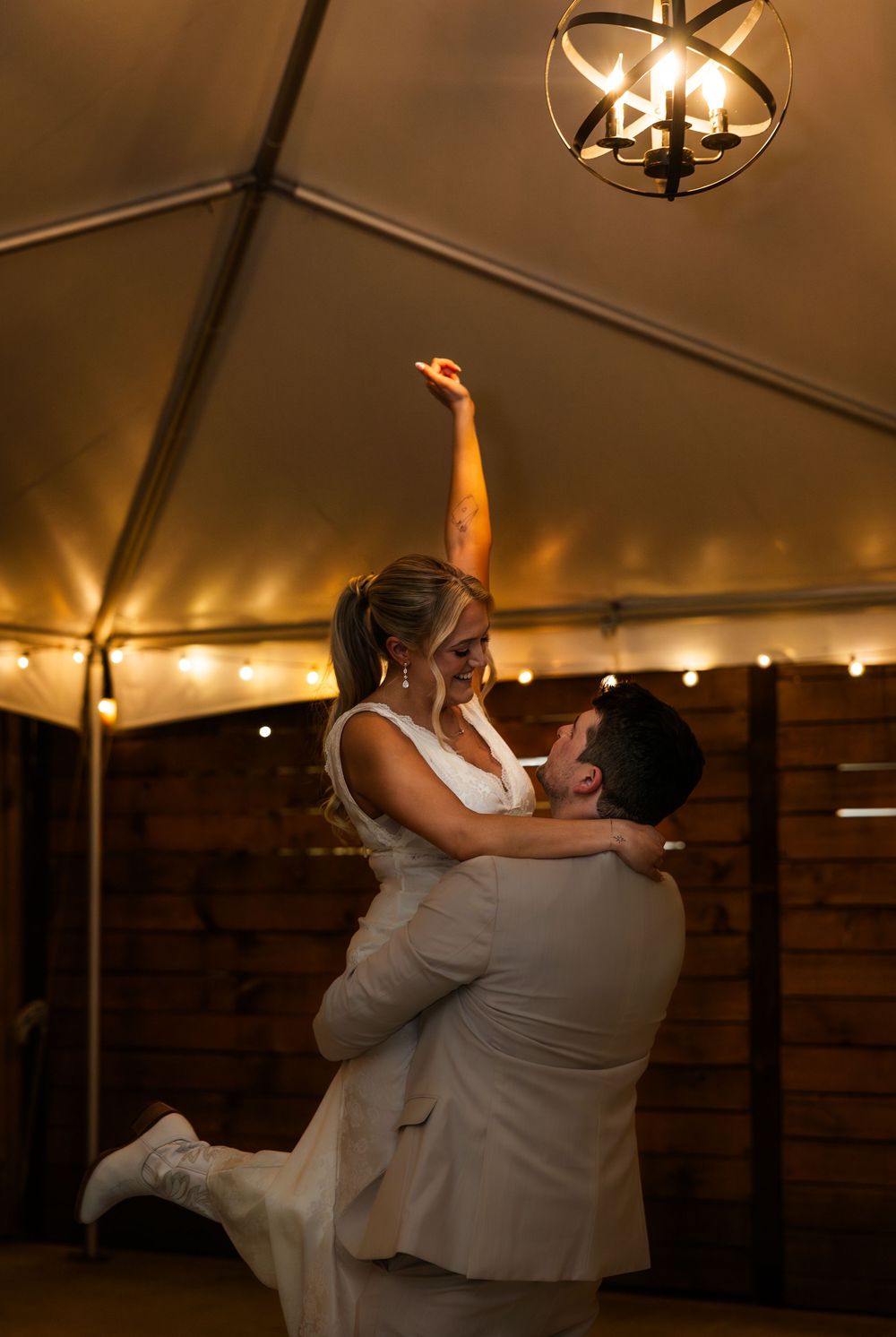 Bride lifted by groom during first dance under warm string lights in elegant tent wedding reception.