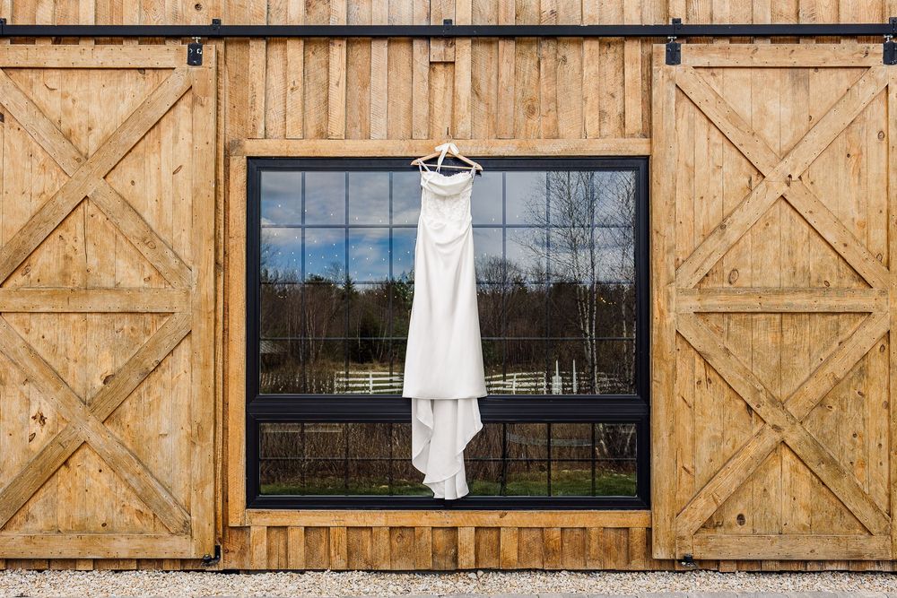 White wedding dress hanging on a rustic wooden barn door with large window reflecting lush greenery outside.