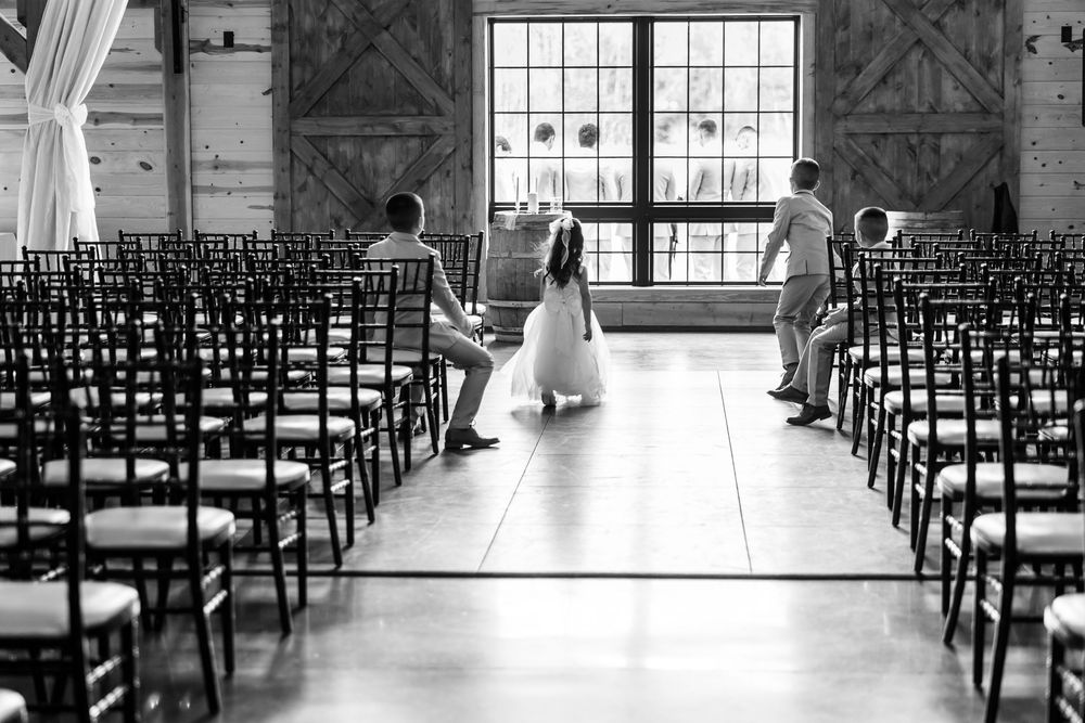 Black and white photo of children playing in a barn wedding venue aisle, surrounded by rows of elegant chiavari chairs.