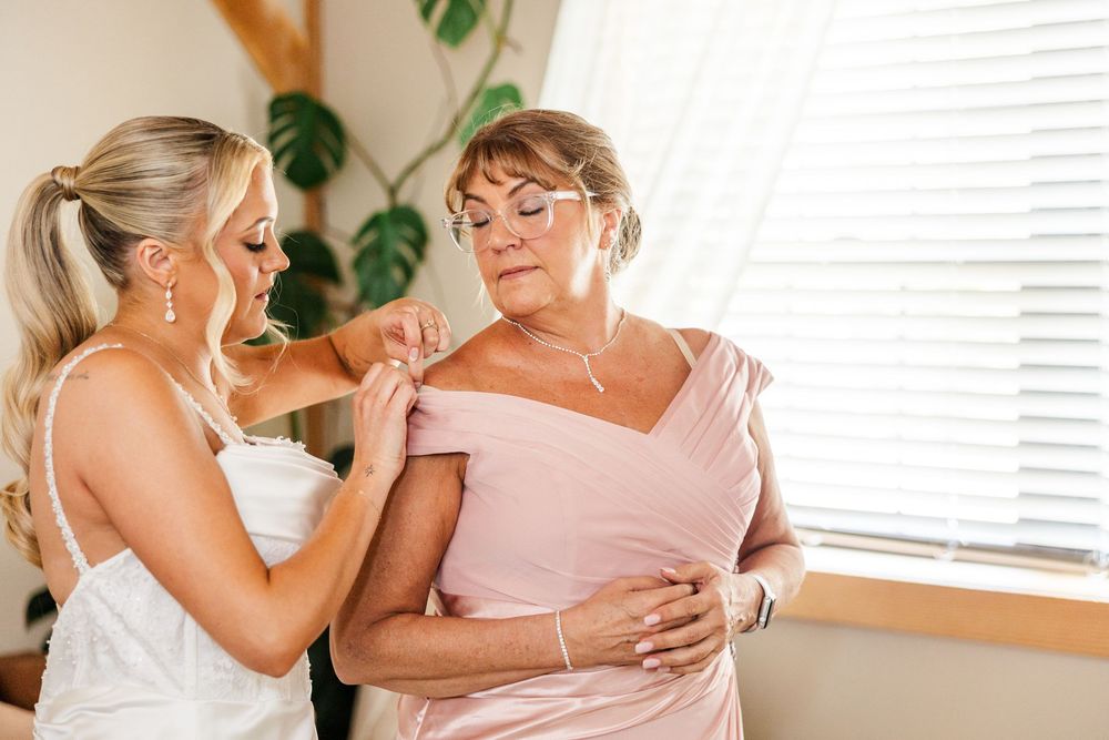 Bride helping older woman in pink dress with shoulder detail while getting ready near bright window.