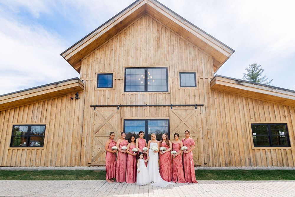 Bride and bridesmaids in pink dresses pose in front of a rustic wooden barn wedding venue.