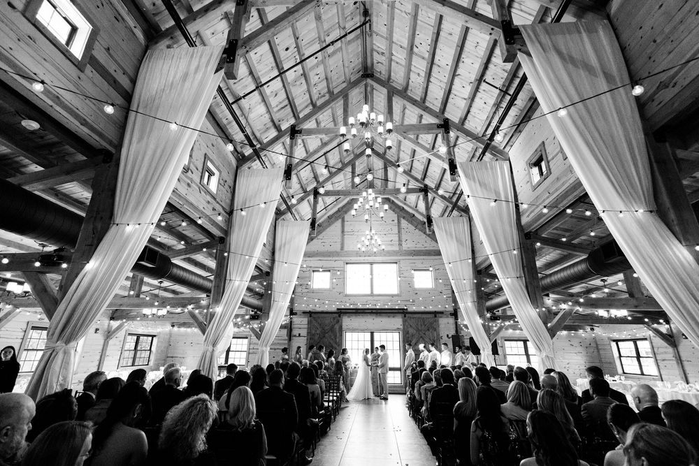 Black and white photo of an elegant barn wedding ceremony with draped fabric, chandeliers, and guests seated in rustic venue.