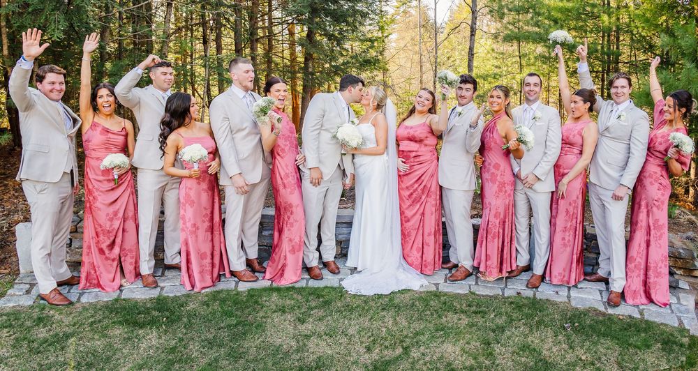 Wedding party in coral bridesmaid dresses and tan suits celebrating outdoors among trees.