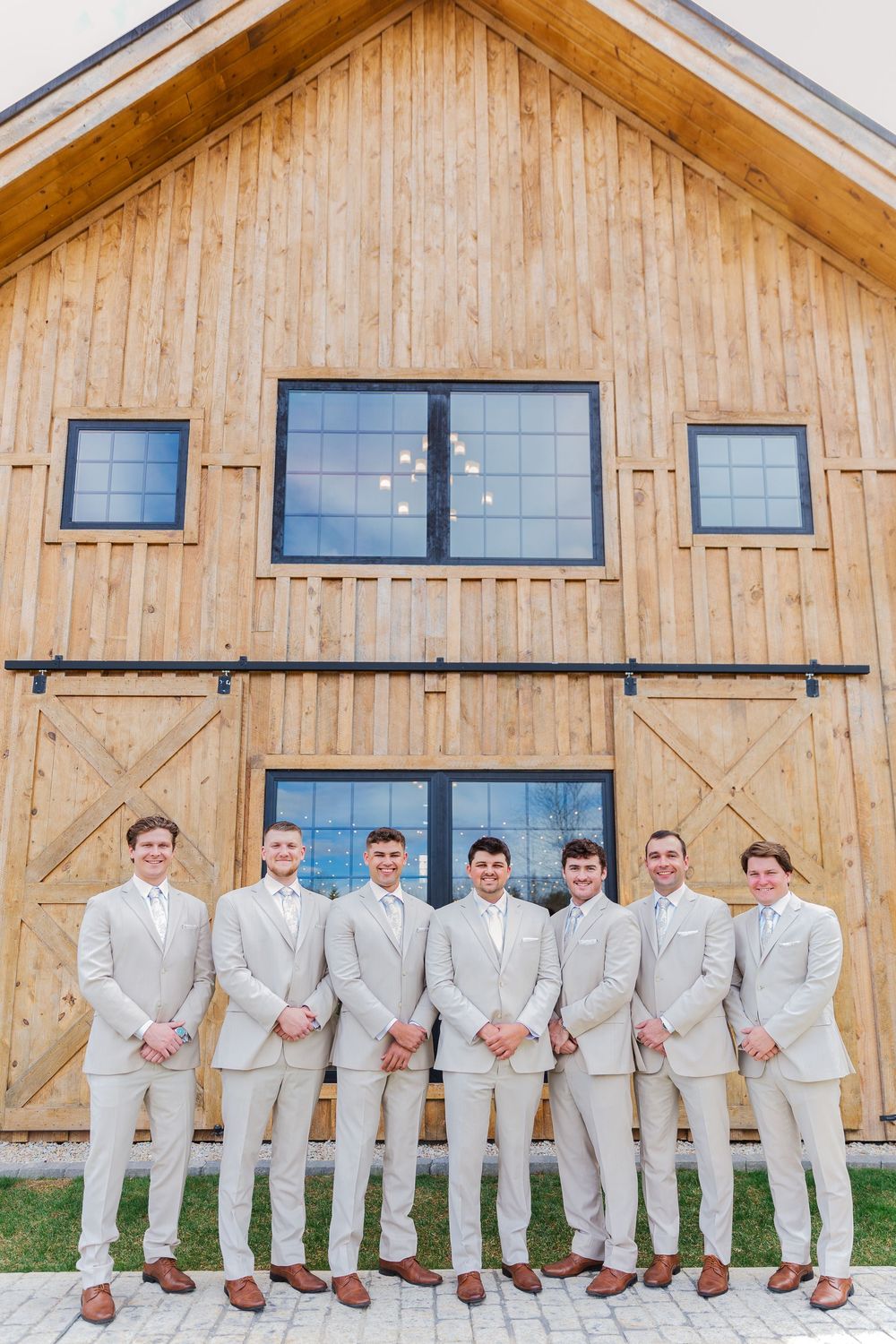 Seven groomsmen in matching light gray suits pose together in front of a rustic wooden barn wedding venue.