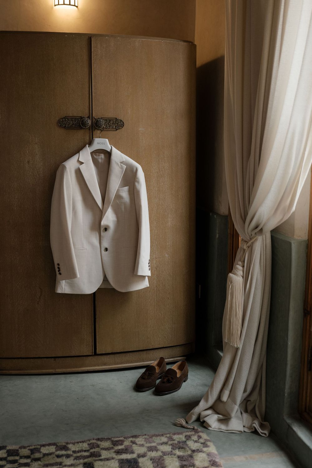 A white suit jacket hangs on a wall in a dimly lit room with checkered floor tiles and flowing curtains.