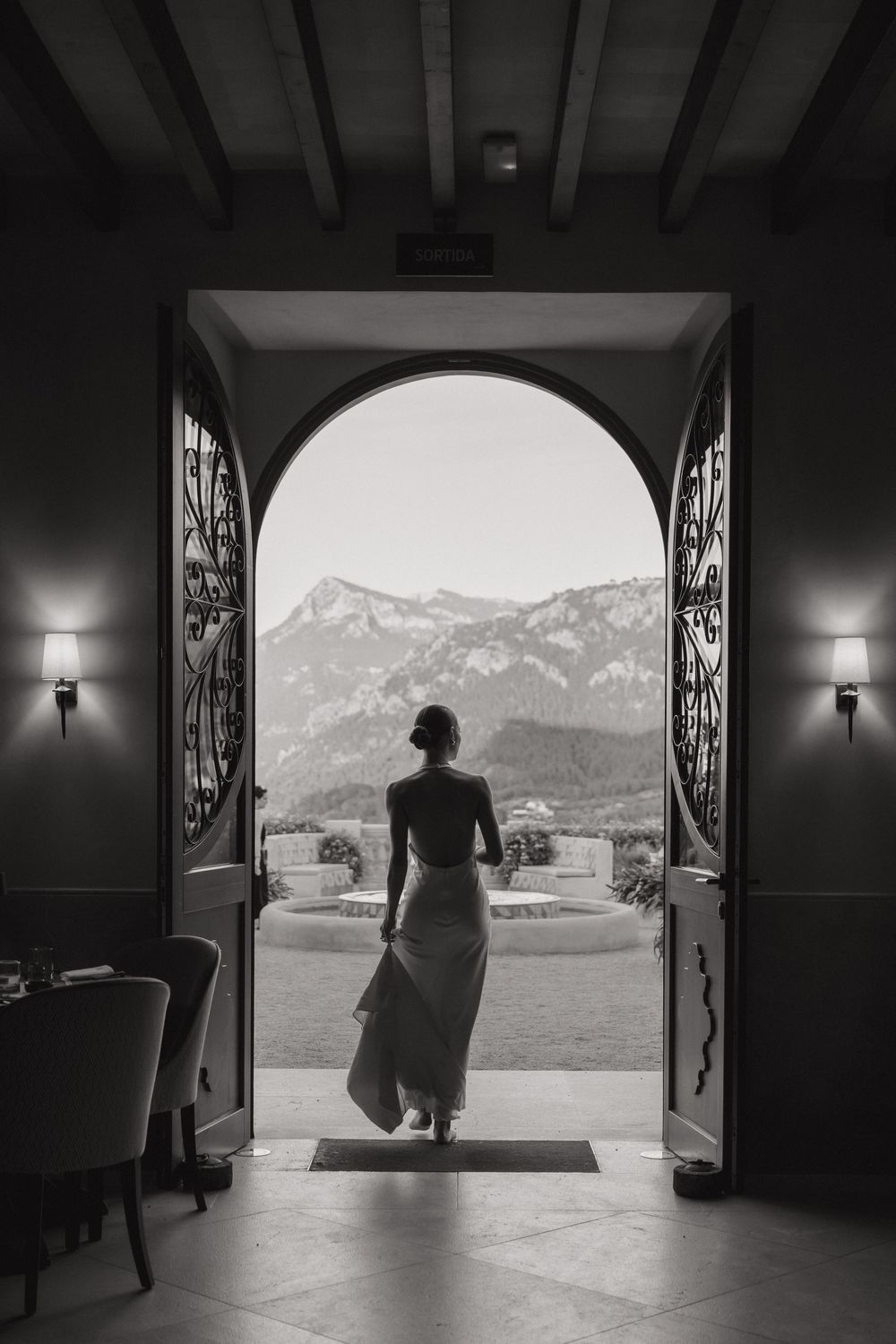 A silhouetted figure stands in a doorway framing a dramatic mountain landscape view in black and white.