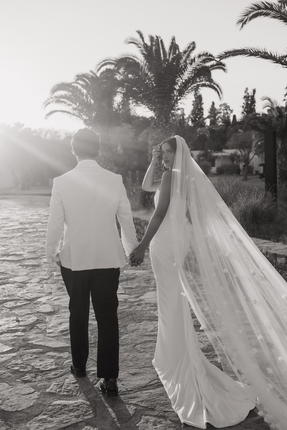 A black and white wedding photo of a couple standing on stone path with palm trees and sunlight behind them.