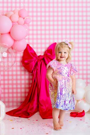 Sparkly pink and purple party dress against gingham background with pink balloons and large red bow decoration. First Birthday Portrait NJ