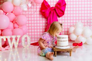 Pink white birthday party setup with balloons, gingham backdrop, and a white decorated cake on a wooden stand. NJ Cake Smash Photographer