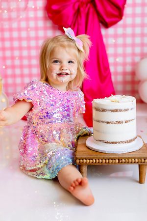 Sparkly sequined dress and white hair bow beside a naked cake on wooden stand against pink gingham backdrop. NJ Cake Smash Photographer