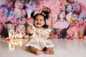 Smiling baby in white lace dress sitting against pink floral backdrop with glowing neon 'One' sign. Cake Smash Photographer in NJ