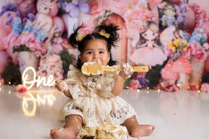 A small child in a lace dress enjoys smashing a cake during a cake smash photo session with floral backdrop and neon 'One' sign. New Jersey