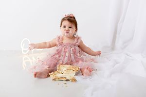 Baby in pink dress sitting on white backdrop with gold decorative elements and crown.