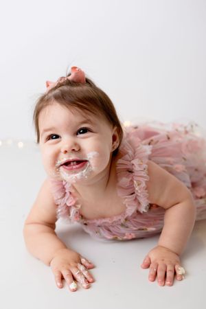 Baby wearing pink tulle dress lies on white background with frosting or cream on face and hands.