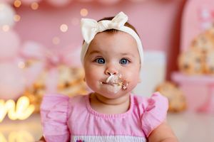 Baby wearing pink dress and white headband sits among soft pink and gold bokeh lighting effects for birthday portraits.