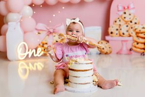 Pink and white first birthday cake smash setup with balloons, neon sign, and cookies in soft pastel colors.