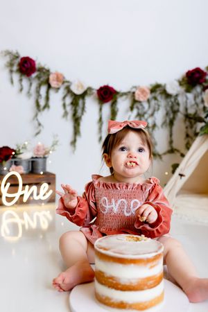 Baby wearing pink dress and bow sits with naked cake decorated with roses for first birthday cake smash photo session.