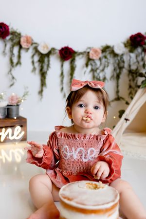A baby wearing a pink smocked dress and floral bow sits at a table with a decorated cake and neon sign.