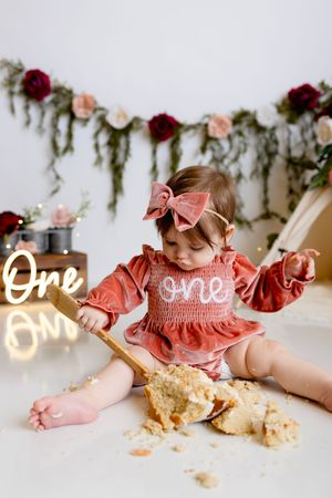 First birthday cake smash photo with coral dress and floral garland backdrop featuring neon 'one' light sign.