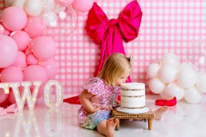 Pink and white first birthday party setup with balloons, gingham pattern backdrop, and cake smash decor.