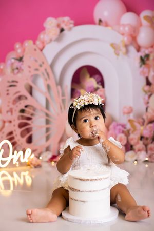 Baby in white dress sits with birthday cake against pink backdrop with balloons and decorative arch for first birthday shoot.