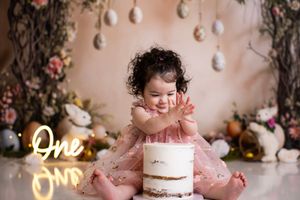 A pink and white cake smash photo session setup with floral decorations and pearls against a beige backdrop.