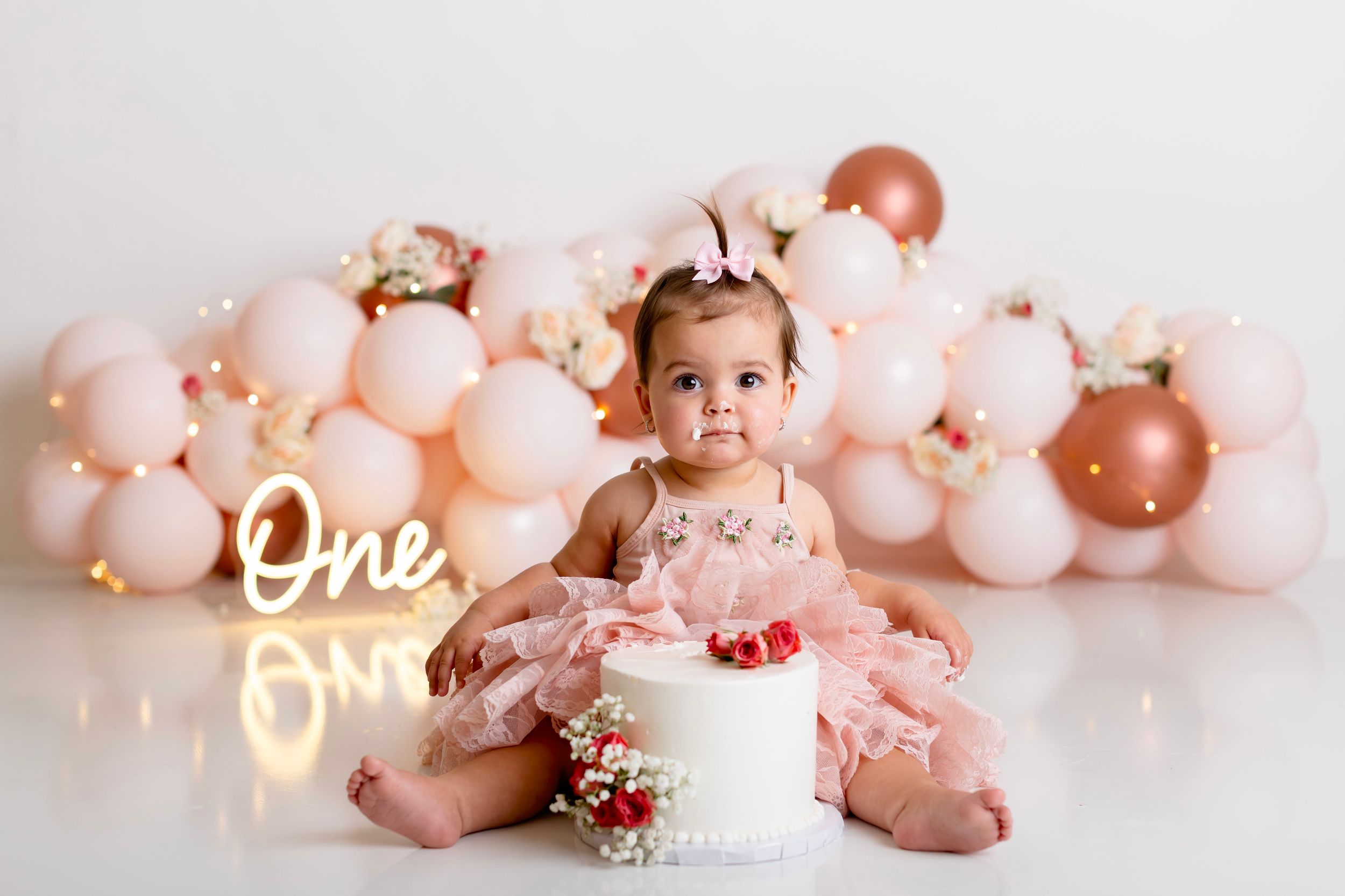 A baby's first birthday photoshoot with pink and rose gold balloons, white cake, and floral decorations.