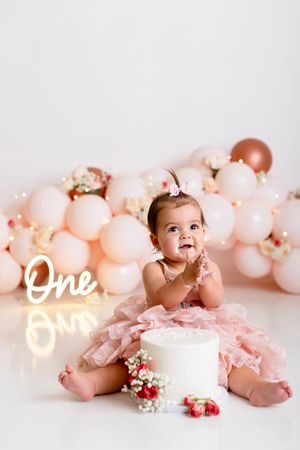 Baby in pink tutu dress lying on floor with pink balloons and floral decorations for first birthday photo shoot.