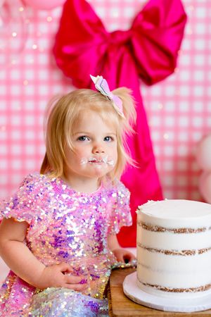 A child in a sparkly pink sequin dress sits next to a white naked cake with a large pink bow backdrop.
