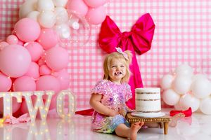 Pink and white balloon arch decorates a birthday cake smash photo session with gingham background.
